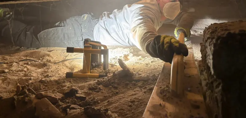 Medewerker van PuinVrij ligt in een kruipruimte en trekt de zandbodem vlak, met een bouwlamp naast zich